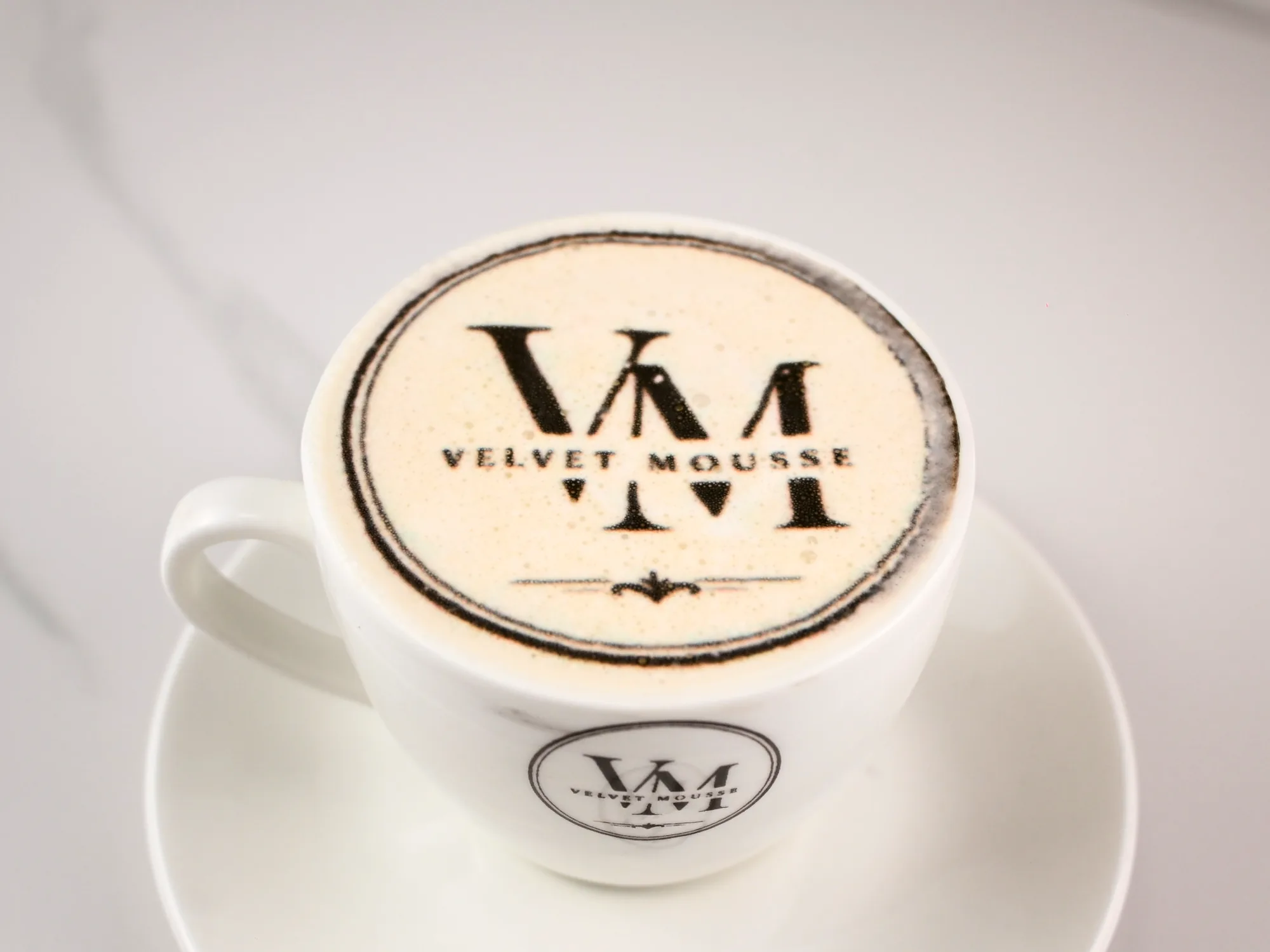 A latte with the Velvet Mousse monogram printed on the foam, in a branded cup and saucer