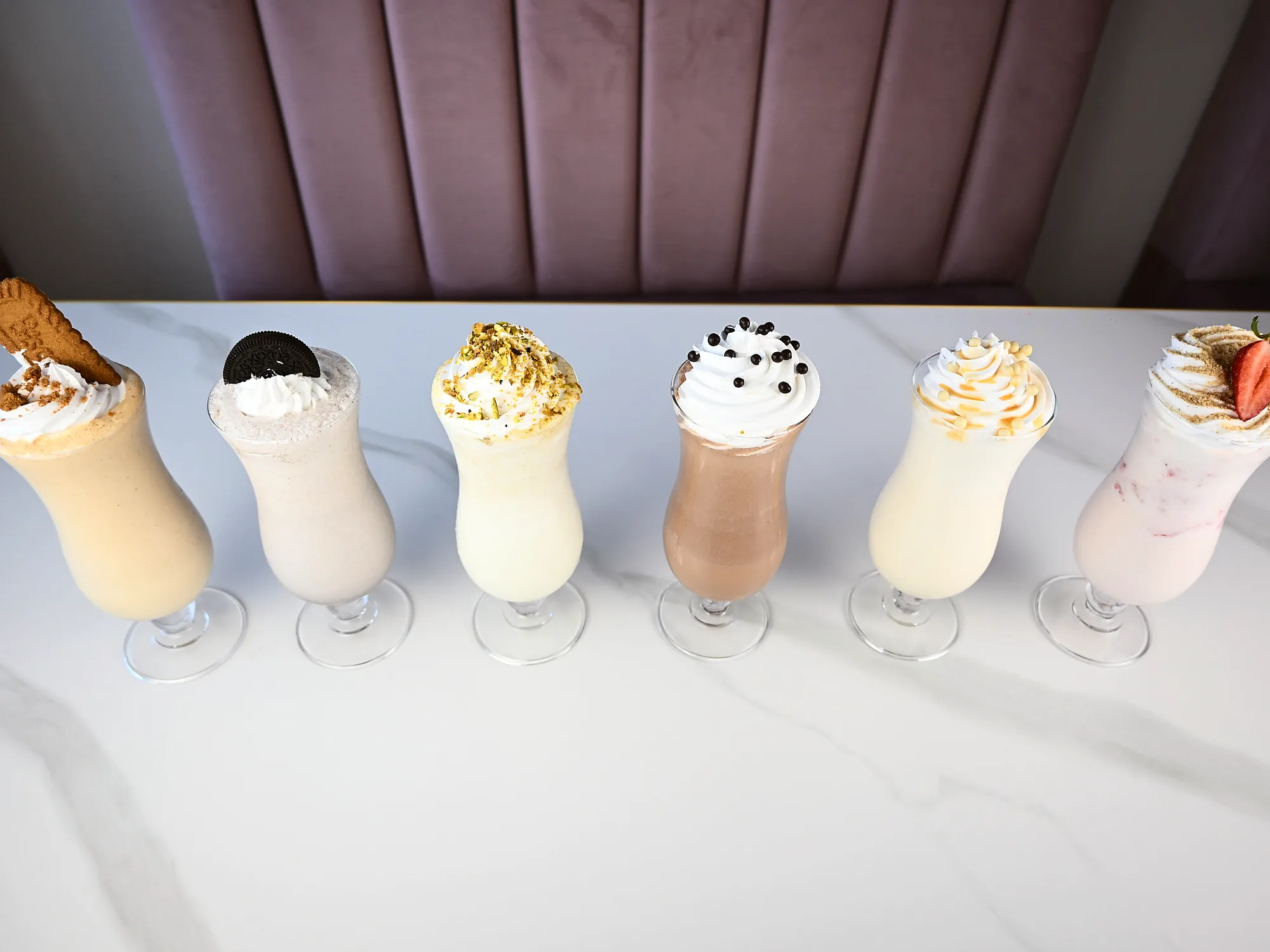 Six milkshakes lined up along a marble counter in front of the pink banquette