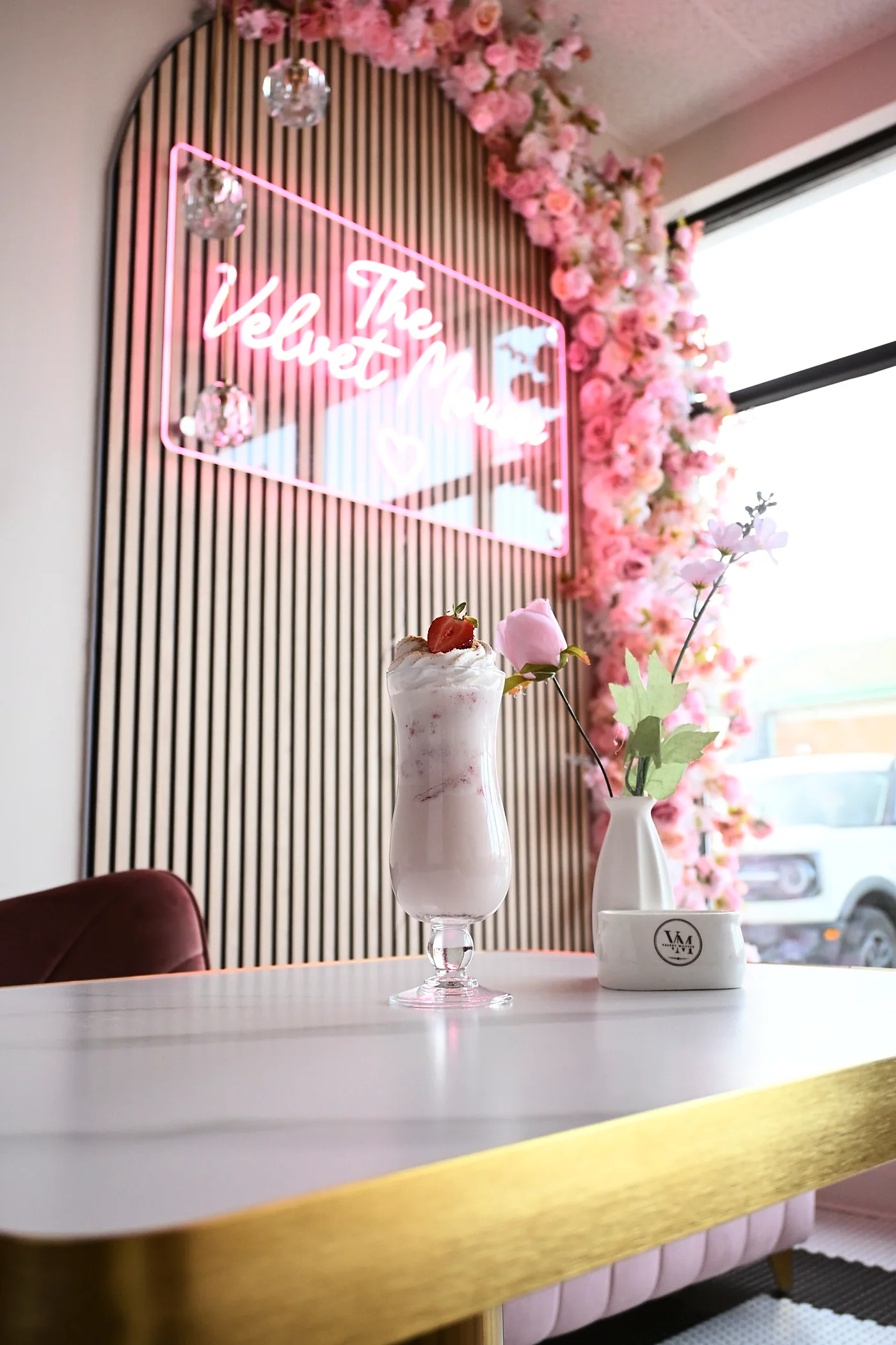 The café's neon Velvet Mousse sign and blossom arch behind a strawberry milkshake on a marble table