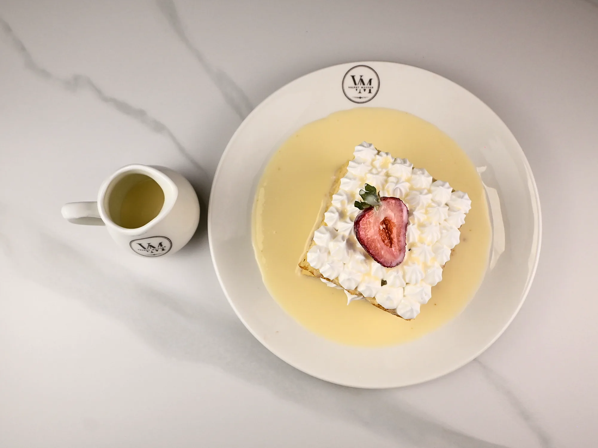 A square of tres leches piped with cream and topped with strawberry, seen from above with a milk jug