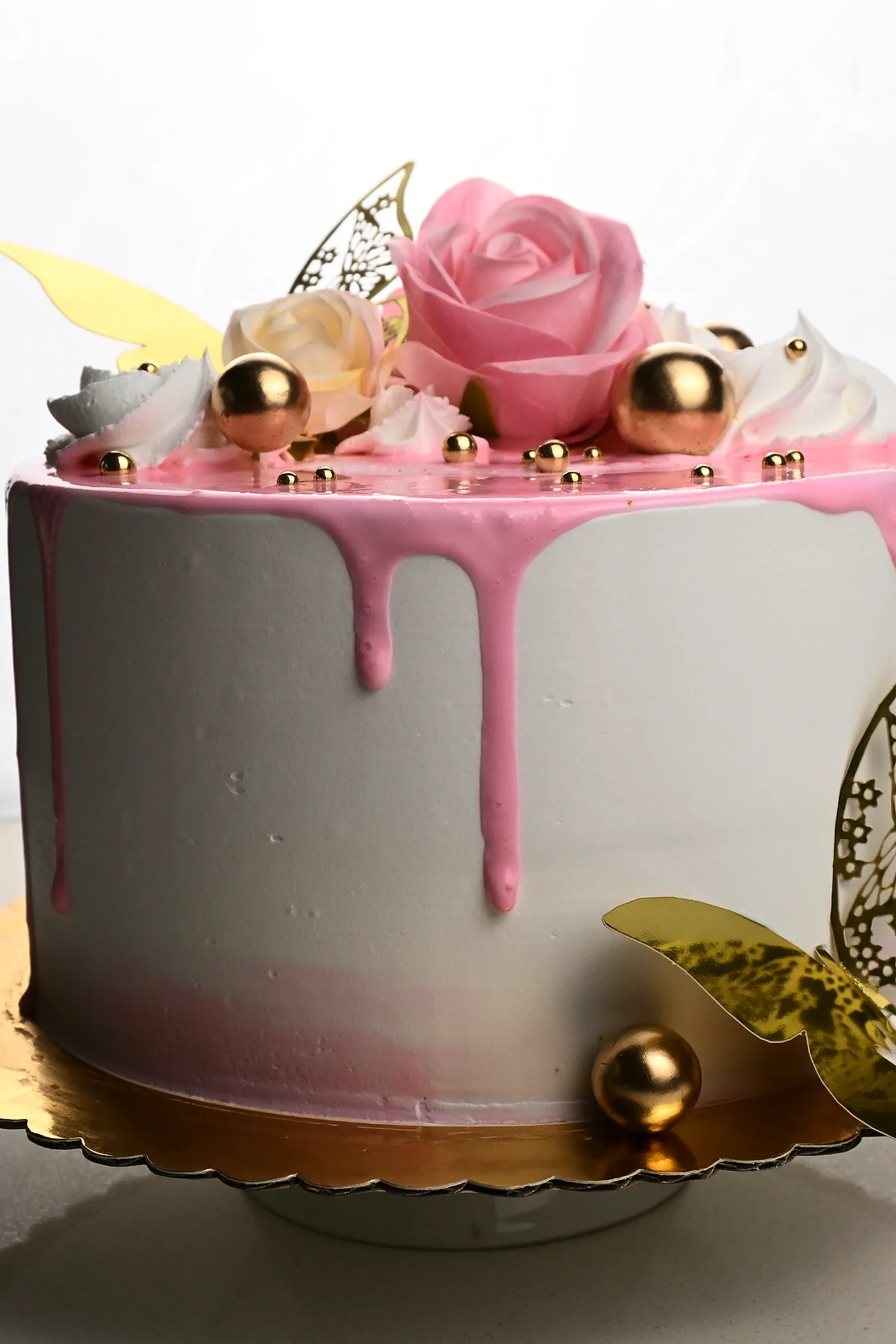 A custom celebration cake with a pink drip, sugar roses, gold spheres and a lace butterfly