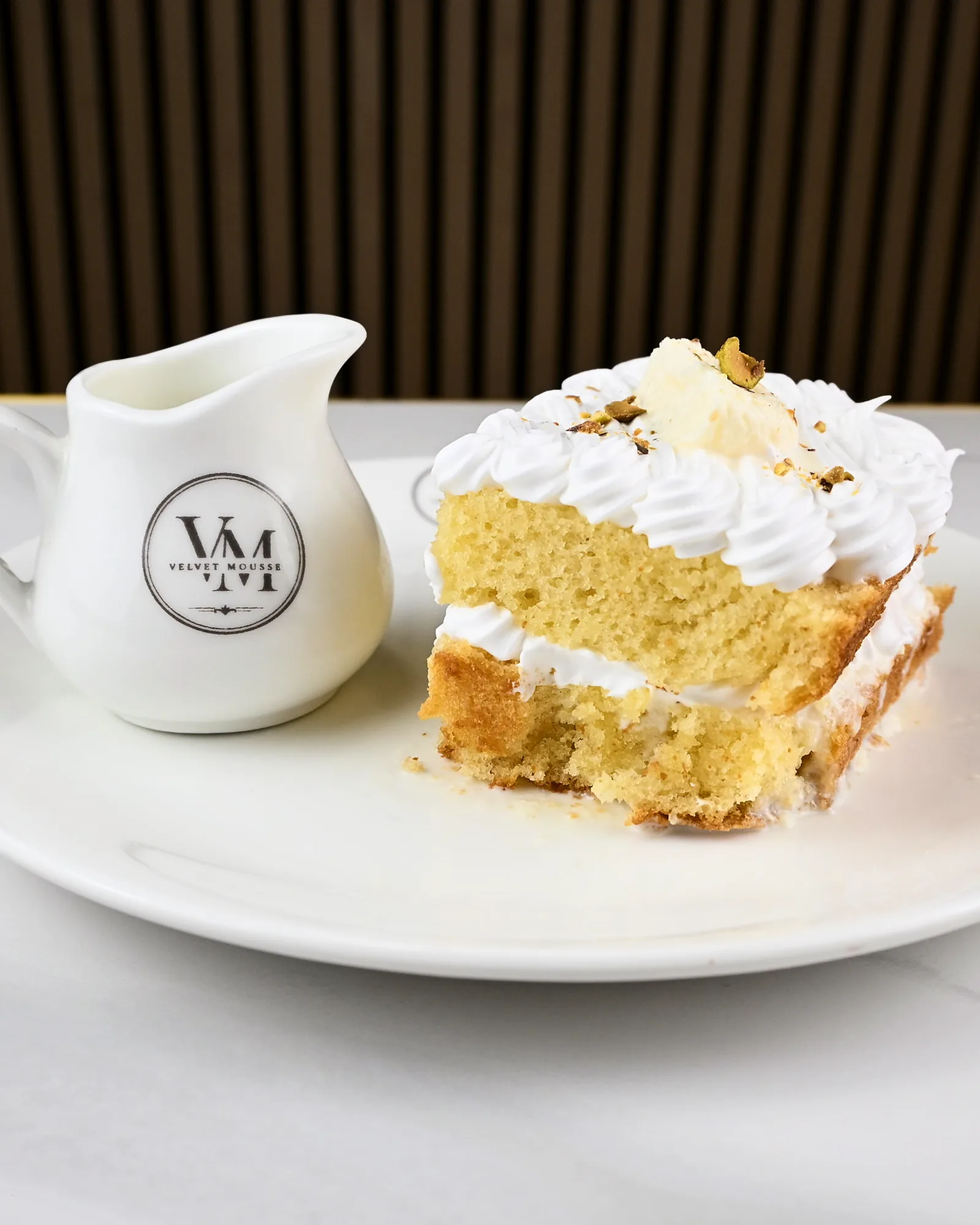 A slice of tres leches with piped cream and a strawberry, beside a milk jug
